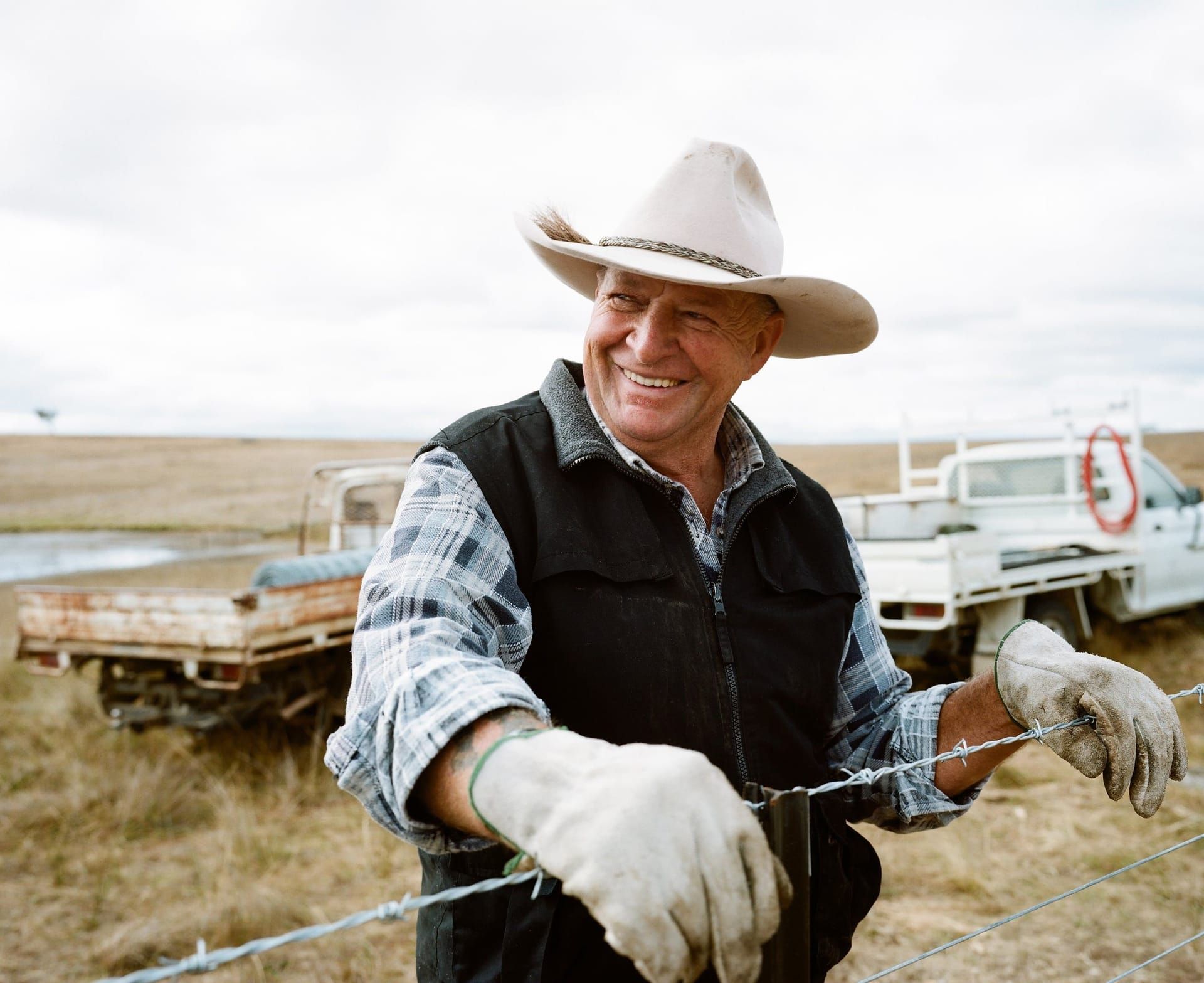 A rancher in a cowboy hat smiling at a barbed-wire fence in the Mountain West.